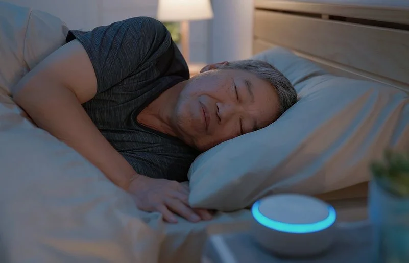 Ein Mann liegt im Bett und schläft. Auf dem Nachttisch neben ihm steht eine kleine, kreisrunde „White-Noise-Machine”.