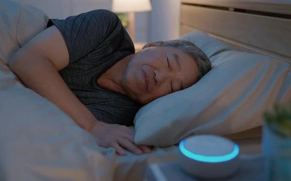 Ein Mann liegt im Bett und schläft. Auf dem Nachttisch neben ihm steht eine kleine, kreisrunde „White-Noise-Machine”.