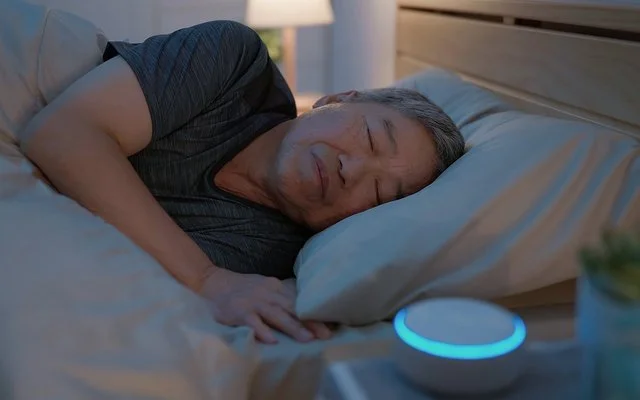 Ein Mann liegt im Bett und schläft. Auf dem Nachttisch neben ihm steht eine kleine, kreisrunde „White-Noise-Machine”.
