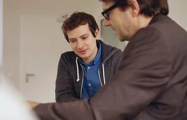 Zwei Personen schauen auf Unterlagen auf einem Tisch. Im Hintergrund erkennt man eine Tür.