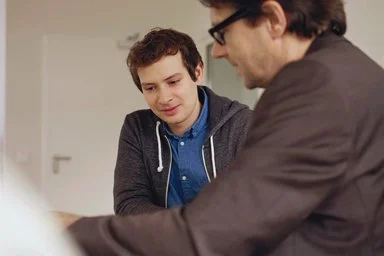 Zwei Personen schauen auf Unterlagen auf einem Tisch. Im Hintergrund erkennt man eine Tür.