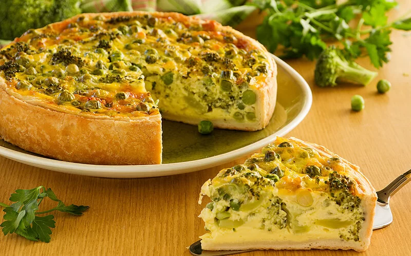 Auf einem Tisch steht ein Gemüsekuchen. Er ist mit Erbsen, Brokkoli und Käse zubereitet. Ein Stückchen ist bereits herausgeschnitten und liegt auf einem Tortenheber.