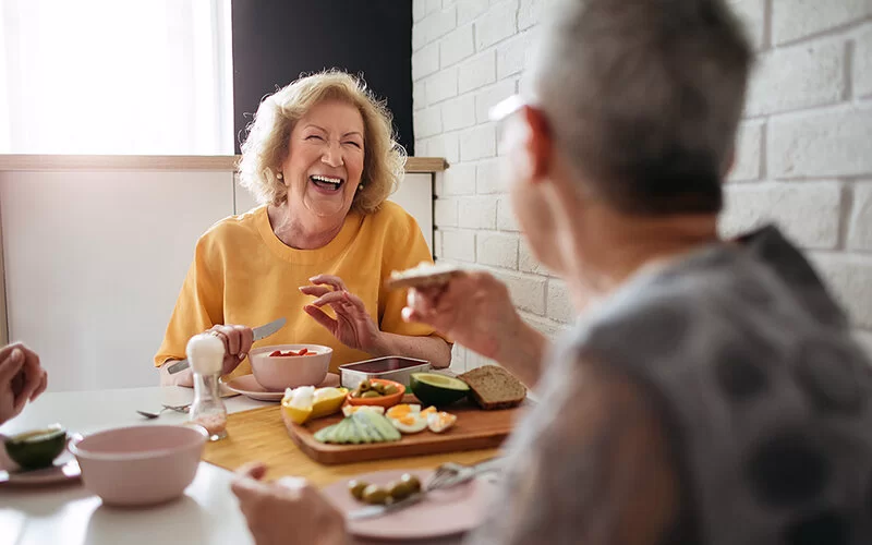 Zwei ältere Frauen sitzen gemeinsam am Frühstückstisch und lachen miteinander.