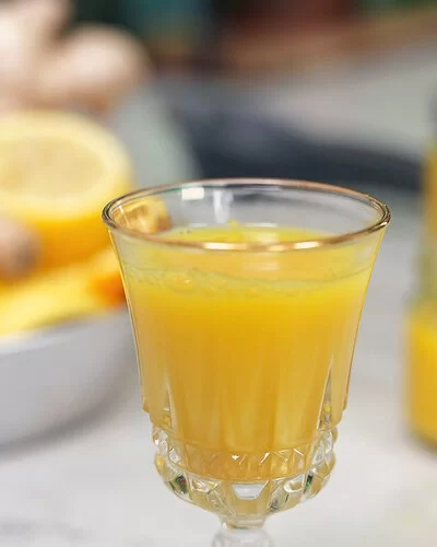 Ingwer-Shot nach einem Rezept von Felicitas Then, serviert in einem Kristallglas. Rechts im Hintergrund weitere Shots in Schraubgläsern. Links im Hintergrund eine silberne Schale voller Ingwerknollen, Zitronen und Orangen.