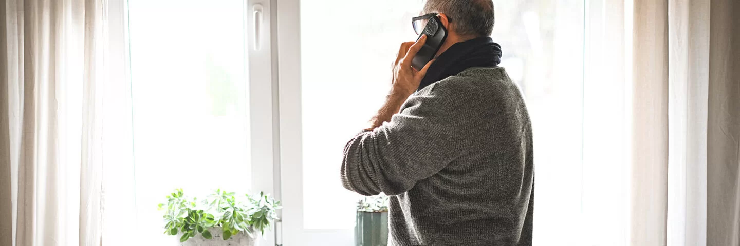 Ein Mann steht am Fenster neben der Heizung und telefoniert. Er spricht über Heizen im Winter.