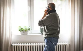 Ein Mann steht am Fenster neben der Heizung und telefoniert. Er spricht über Heizen im Winter.
