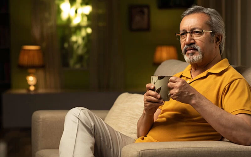 Ein älterer Mann sitzt auf der Couch und trinkt vor dem Schlafengehen einen Tee als entspannendes Ritual.