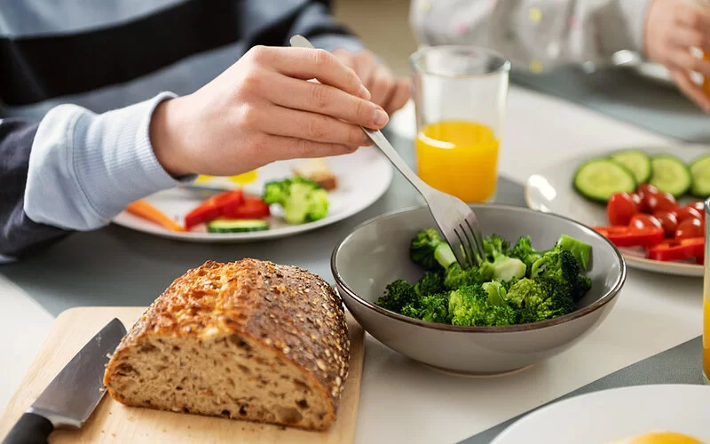 Ein mit Gemüse und Vollkornbrot gedeckter Tisch. Eine Hand pickt mit einer Gabel Brokkoli aus einer Schüssel.