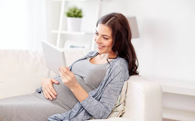 Eine junge Frau liegt auf dem Sofa und lehnt sich mit dem Rücken an die Armlehne. Mit der rechten Hand fasst sie sich an den Bauch, in der linken hält sie ein Tablet. Sie betrachtet den Bildschirm und lächelt dabei.