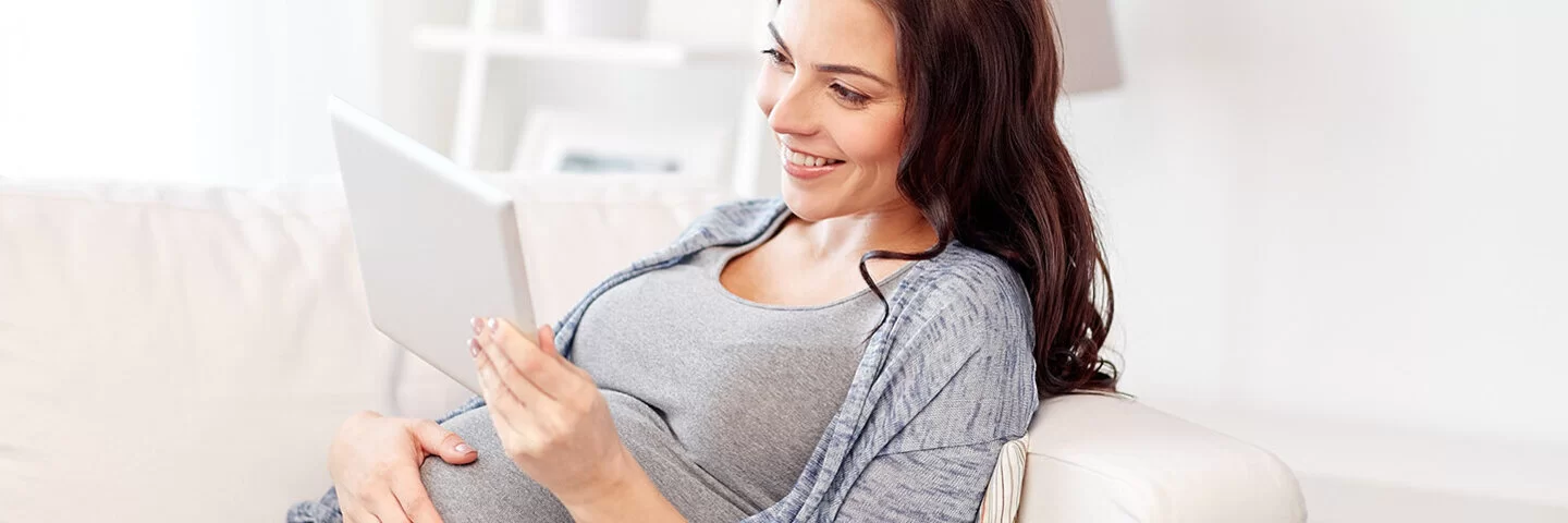 Eine junge Frau liegt auf dem Sofa und lehnt sich mit dem Rücken an die Armlehne. Mit der rechten Hand fasst sie sich an den Bauch, in der linken hält sie ein Tablet. Sie betrachtet den Bildschirm und lächelt dabei.