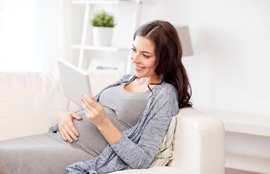 Eine junge Frau liegt auf dem Sofa und lehnt sich mit dem Rücken an die Armlehne. Mit der rechten Hand fasst sie sich an den Bauch, in der linken hält sie ein Tablet. Sie betrachtet den Bildschirm und lächelt dabei.