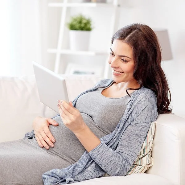 Eine junge Frau liegt auf dem Sofa und lehnt sich mit dem Rücken an die Armlehne. Mit der rechten Hand fasst sie sich an den Bauch, in der linken hält sie ein Tablet. Sie betrachtet den Bildschirm und lächelt dabei.