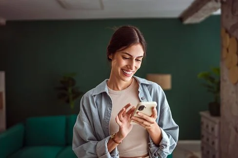 Eine junge Frau steht in einem gemütlich eingerichteten Wohnzimmer und hält ein Smartphone in der linken Hand, mit den Fingern ihrer rechten Hand tippt sie auf das Display. Sie lächelt beim Lesen ihrer Nachrichten.