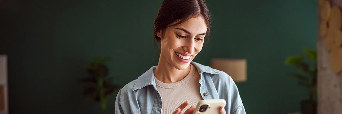 Eine junge Frau steht in einem gemütlich eingerichteten Wohnzimmer und hält ein Smartphone in der linken Hand, mit den Fingern ihrer rechten Hand tippt sie auf das Display. Sie lächelt beim Lesen ihrer Nachrichten.