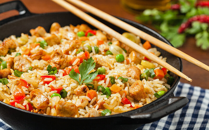 Auf einem blau-weiß karierten Küchentuch steht ein gusseiserner Wok mit gebratenem Gemüse-Reis und Hähnchen-Stückchen. Darauf liegt ein Paar Essstäbchen.