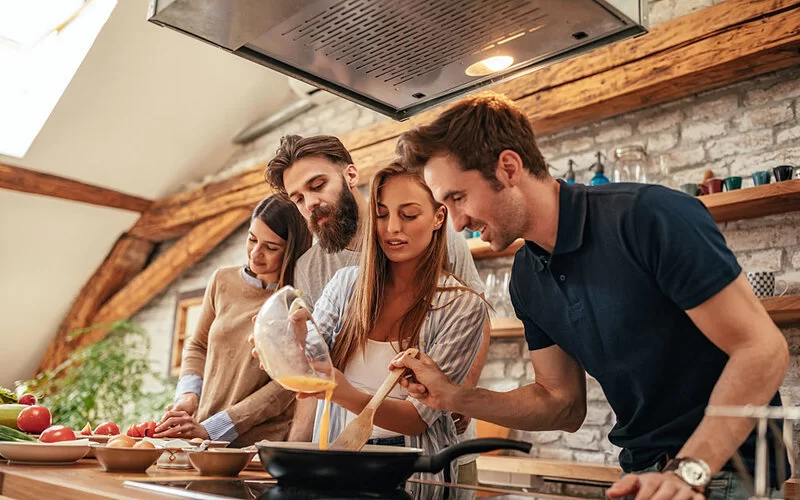 Zwei junge Männer und zwei junge Frauen stehen an einer Kücheninsel und kochen gemeinsam.