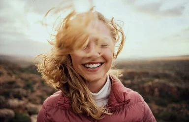 Eine Frau mit blonden Haaren, die im Wind wehen, und einer rosafarbenen Steppjacke steht lächelnd und mit geschlossenen Augen inmitten einer Landschaft.