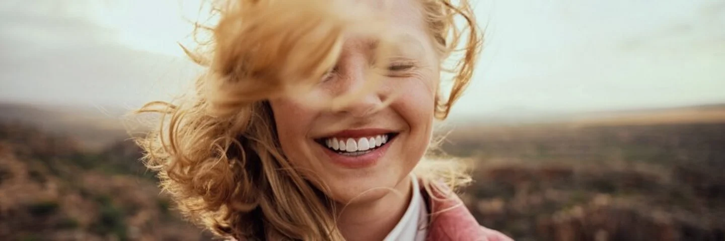 Eine Frau mit blonden Haaren, die im Wind wehen, und einer rosafarbenen Steppjacke steht lächelnd und mit geschlossenen Augen inmitten einer Landschaft.