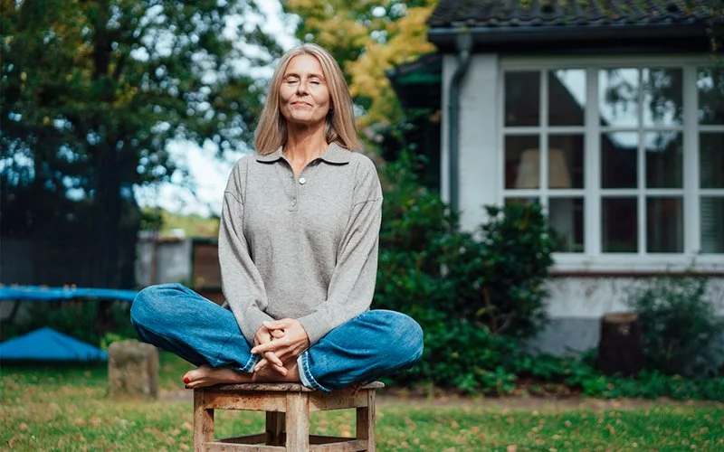 Frau sitzt entspannt im Schneidersitz auf einem Hocker im Garten vor einem Haus; Hände locker auf den Knien, gelassener Blick.
