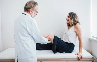 In einer Arztpraxis sitzt eine Frau auf einer Liege. Der Arzt hat ihr linkes Bein angewinkelt und tastet die Kniekehle ab.