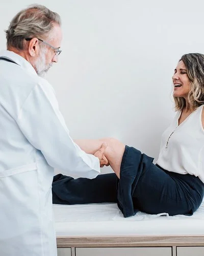 In einer Arztpraxis sitzt eine Frau auf einer Liege. Der Arzt hat ihr linkes Bein angewinkelt und tastet die Kniekehle ab.