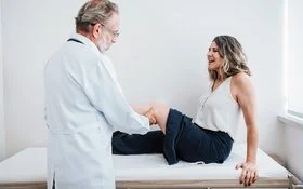 In einer Arztpraxis sitzt eine Frau auf einer Liege. Der Arzt hat ihr linkes Bein angewinkelt und tastet die Kniekehle ab.