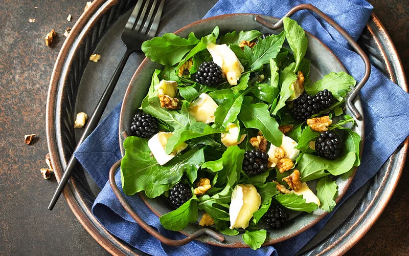 Ein Rucola-Salat mit Brombeer-Vinaigrette, frischen Früchten, Nüssen und Weichkäse. Schale auf einer Steinarbeitsplatte.