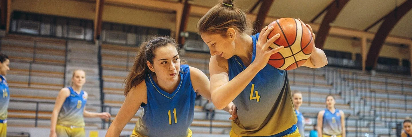 Eine Frauenbasketballmannschaft trainiert in einer Sporthalle. Im Vordergrund steht eine Spielerin, die den Ball hält und von einer zweiten Spielerin gedeckt wird.