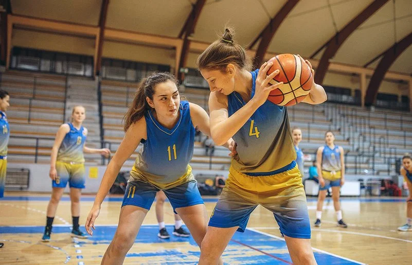 Eine Frauenbasketballmannschaft trainiert in einer Sporthalle. Im Vordergrund steht eine Spielerin, die den Ball hält und von einer zweiten Spielerin gedeckt wird.