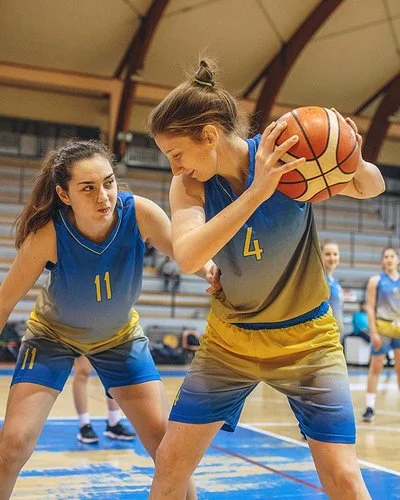 Eine Frauenbasketballmannschaft trainiert in einer Sporthalle. Im Vordergrund steht eine Spielerin, die den Ball hält und von einer zweiten Spielerin gedeckt wird.
