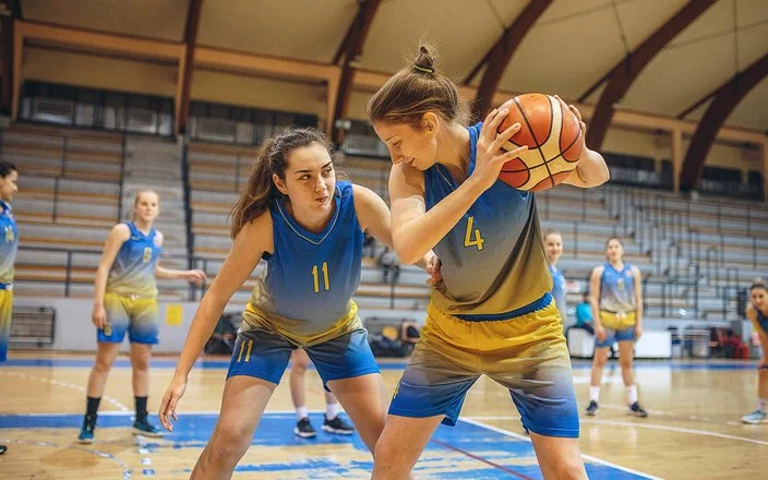 Eine Frauenbasketballmannschaft trainiert in einer Sporthalle. Im Vordergrund steht eine Spielerin, die den Ball hält und von einer zweiten Spielerin gedeckt wird.