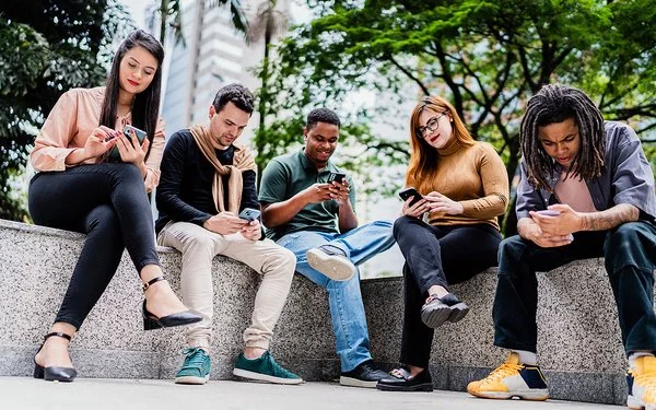 Fünf Freunde oder Co-Worker sitzen nebeneinander auf einer niedrigen Mauer im Park, während jeder auf sein oder ihr Handy schaut.