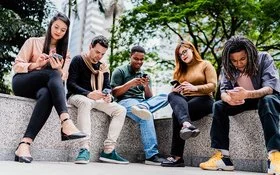 Fünf Freunde oder Co-Worker sitzen nebeneinander auf einer niedrigen Mauer im Park, während jeder auf sein oder ihr Handy schaut.