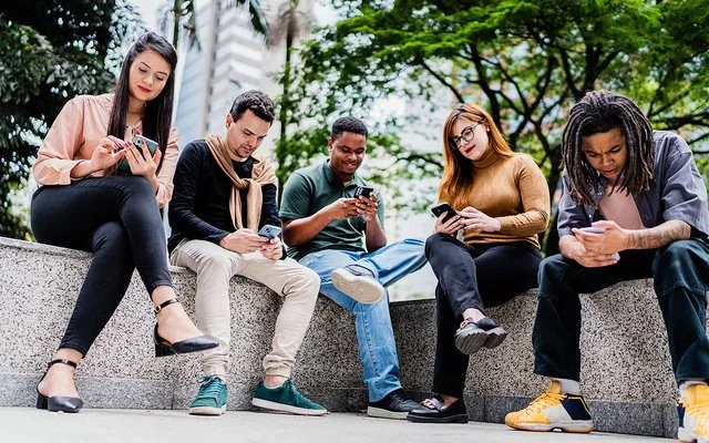 Fünf Freunde oder Co-Worker sitzen nebeneinander auf einer niedrigen Mauer im Park, während jeder auf sein oder ihr Handy schaut.