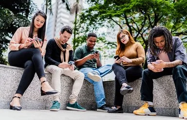 Fünf Freunde oder Co-Worker sitzen nebeneinander auf einer niedrigen Mauer im Park, während jeder auf sein oder ihr Handy schaut.