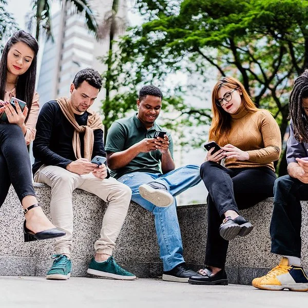 Fünf Freunde oder Co-Worker sitzen nebeneinander auf einer niedrigen Mauer im Park, während jeder auf sein oder ihr Handy schaut.