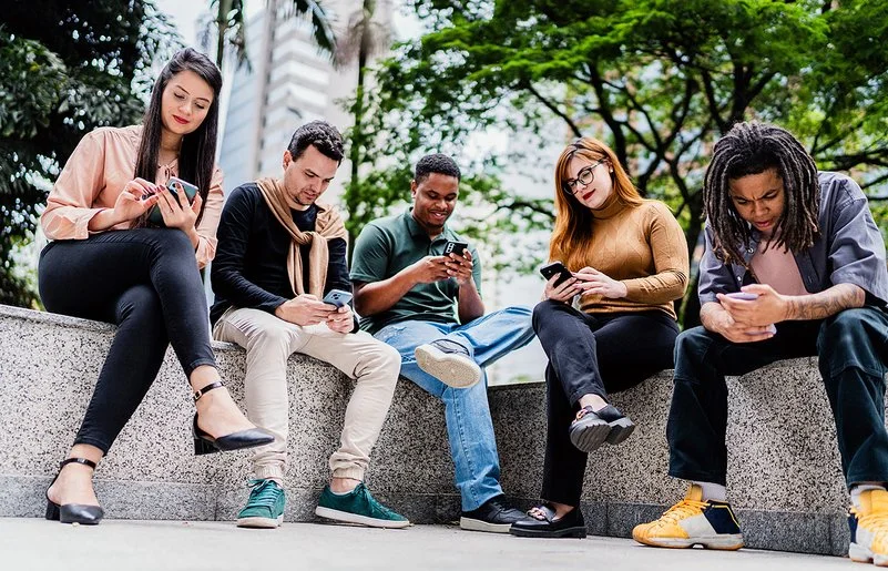 Fünf Freunde oder Co-Worker sitzen nebeneinander auf einer niedrigen Mauer im Park, während jeder auf sein oder ihr Handy schaut.