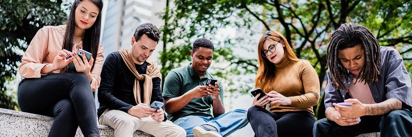 Fünf Freunde oder Co-Worker sitzen nebeneinander auf einer niedrigen Mauer im Park, während jeder auf sein oder ihr Handy schaut.