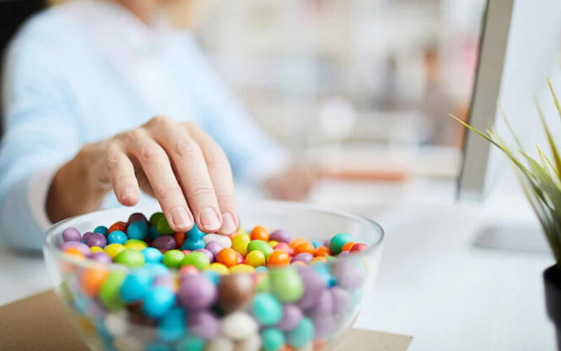 Eine Frau ist süchtig nach Zucker und greift beim Arbeiten in eine Schale mit zuckerhaltigen Schokolinsen.