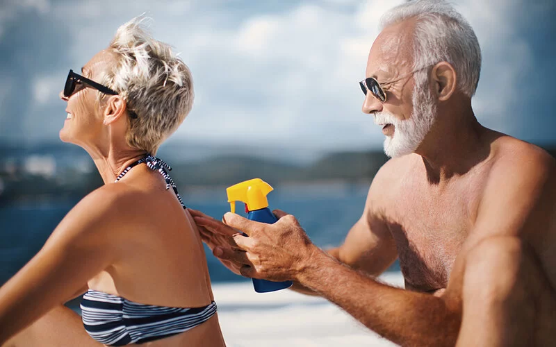 Ein älteres Ehepaar sitzt in Badebekleidung am Strand, er trägt auf ihrem Rücken Sonnencreme auf.