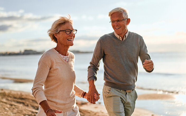 Ein älteres Paar läuft lächelnd Hand in Hand am Strand entlang.