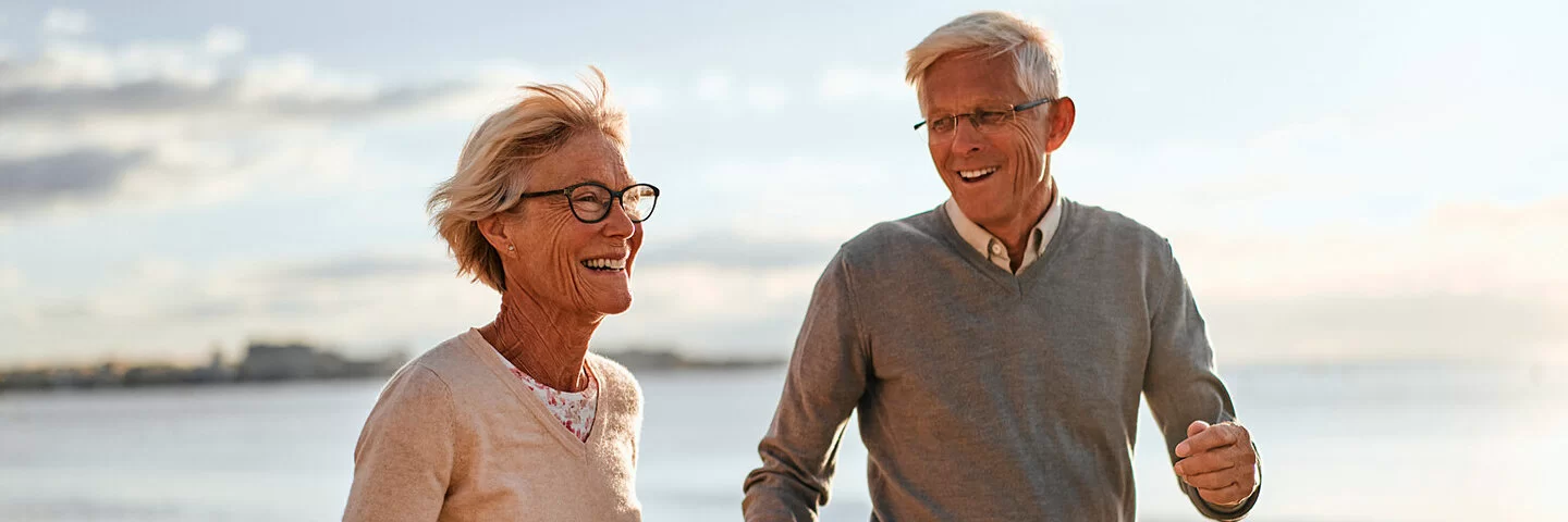 Ein älteres Paar läuft lächelnd Hand in Hand am Strand entlang.