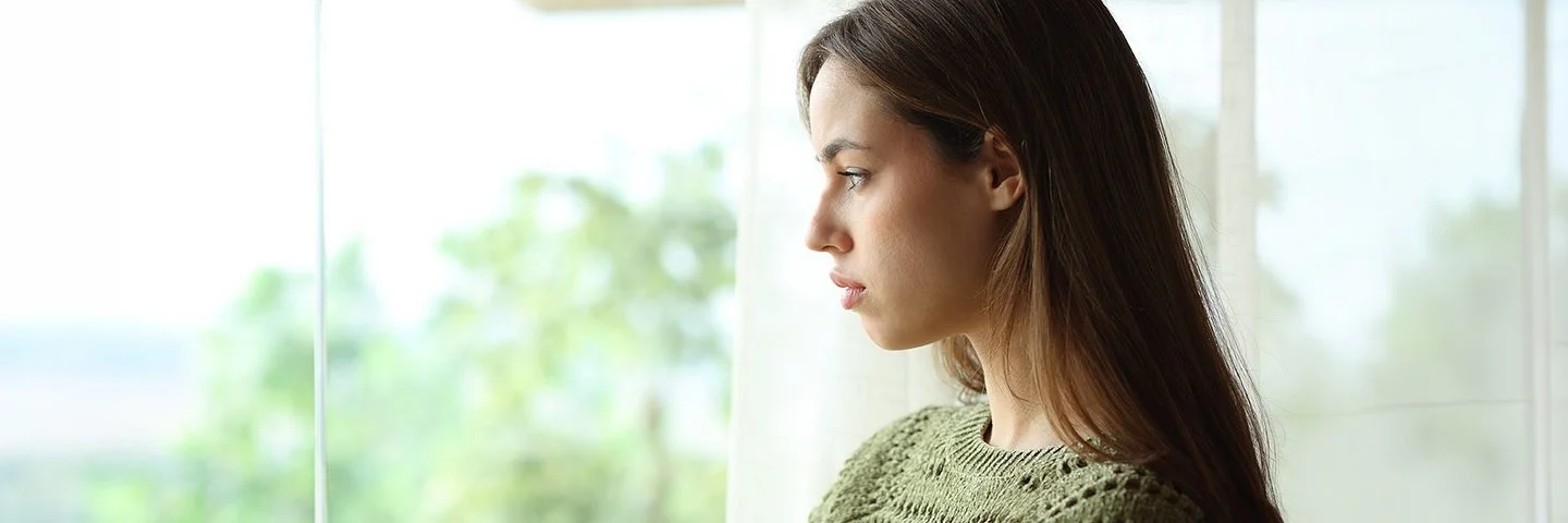Eine junge Frau in einem grünen Pullover schaut traurig aus dem Fenster ins Grüne.
