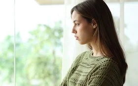 Eine junge Frau in einem grünen Pullover schaut traurig aus dem Fenster ins Grüne.