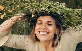 Eine lachende Frau hat die Augen geschlossen und hält eine Blumenkrone auf ihrem Kopf.