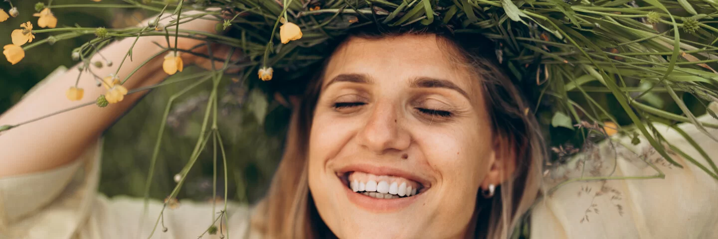 Eine lachende Frau hat die Augen geschlossen und hält eine Blumenkrone auf ihrem Kopf.