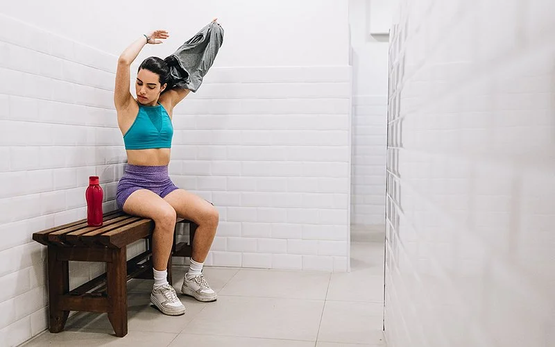 Eine Frau mit sportlicher Figur sitzt auf einer Holzbank in einem Umkleideraum mit weißen Fliesenwänden. Sie trägt ein türkisfarbenes Sportoberteil, eine lilafarbene kurze Hose und weiße Sportschuhe. Sie hebt ein graues Oberteil über den Kopf. Neben ihr steht eine rote Trinkflasche auf der Bank. Die Umgebung wirkt sauber und aufgeräumt.