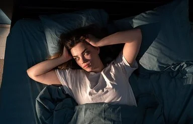 Eine junge Frau liegt mit offenen Augen im Bett und fasst sich mit den Händen an den Kopf.