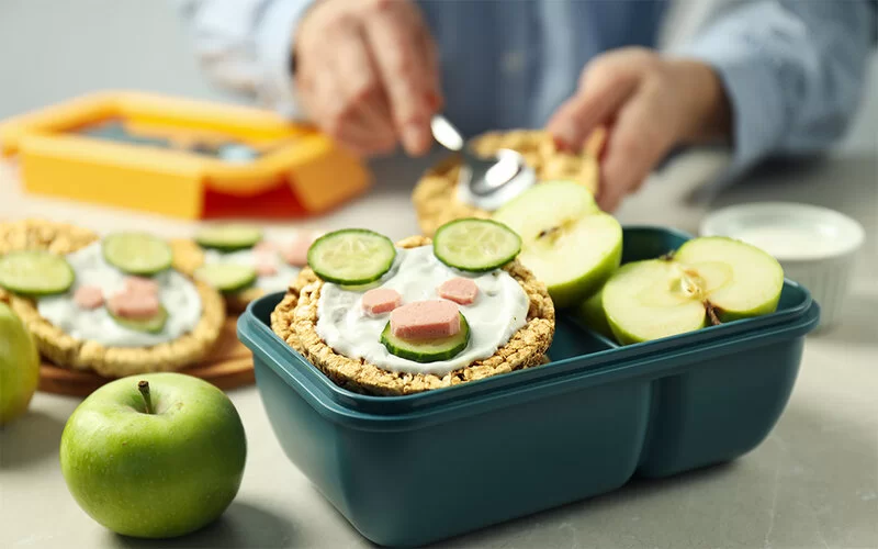 Eine Lunchbox mit belegten Maiswaffeln steht auf einer Küchenplatte. Im Hintergrund bereitet eine Person die Snacks zu.