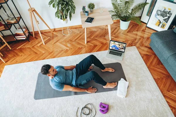 Das Bild zeigt einen jungen Mann im Wohnzimmer auf einer Yogamatte. Neben ihm steht ein Laptop, auf dem ein Workout-Video eingeschaltet ist.